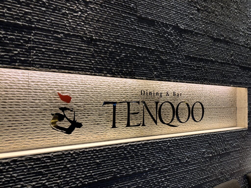 TENQOO店内サイン「ダイニング&バー『TENQOO』の店内壁面サイン。黒いタイル調の壁に白く光るロゴが映える。」