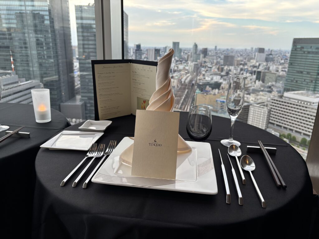 窓際テーブルセッティング「TENQOOの窓際テーブルセッティング。東京の高層ビル群を背景に、白い皿とシルバーのカトラリー、シャンパングラスが並ぶ。」