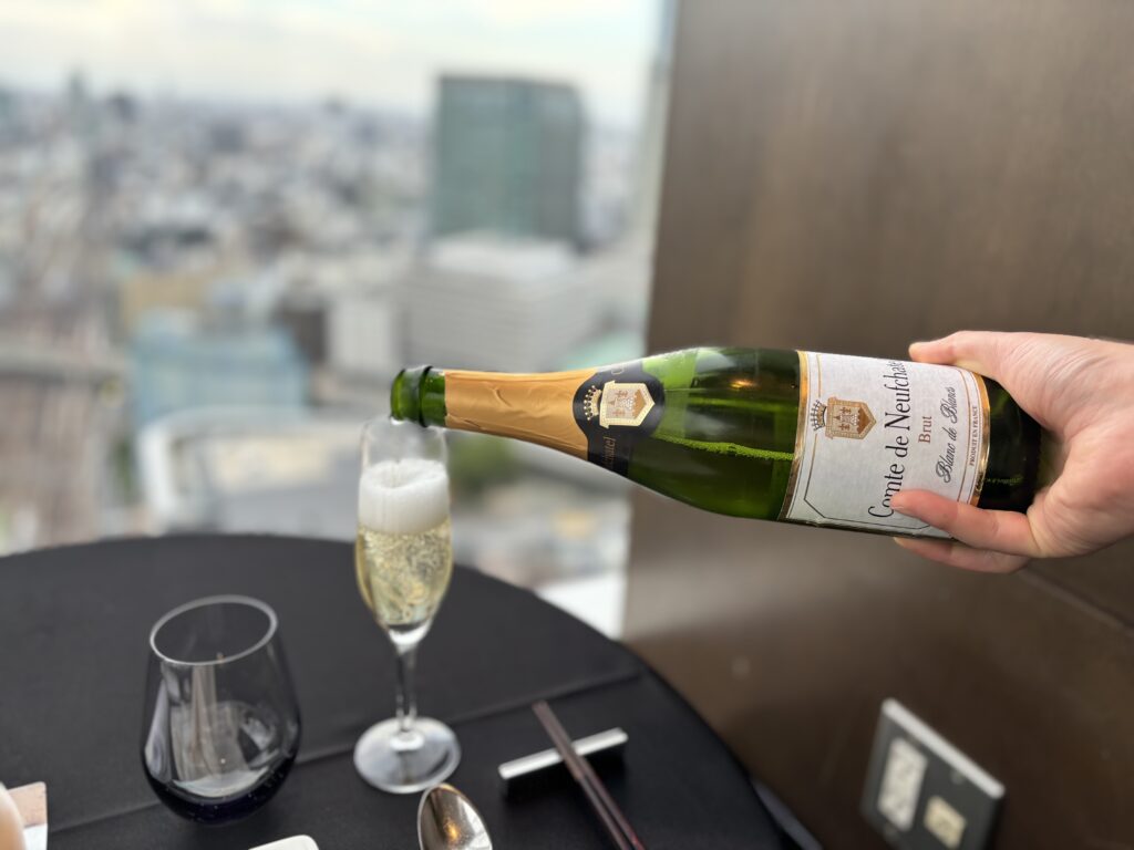 スパークリングワインを注ぐ様「Comte de Neuchatelのスパークリングワインをグラスに注ぐ手元。背景には高層ビル群が広がる。」