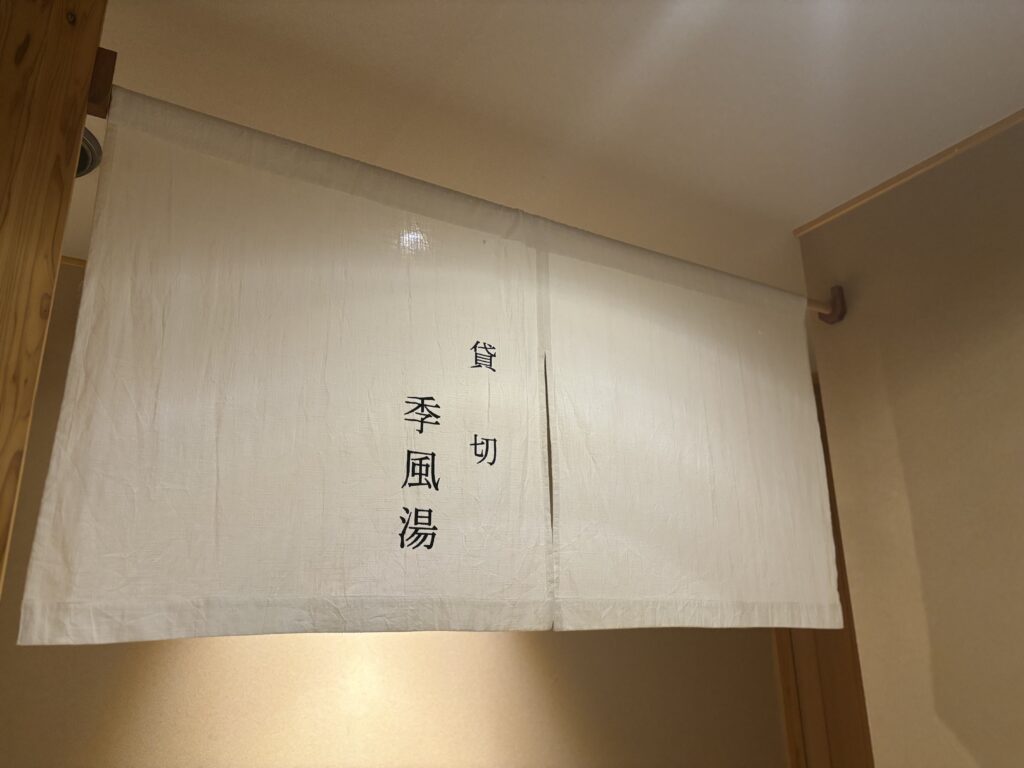 貸切風呂「季風湯」暖簾。吉田屋の貸切風呂「季風湯」の暖簾。白い布にシンプルな文字で温泉の入り口を示す落ち着いた雰囲気。
